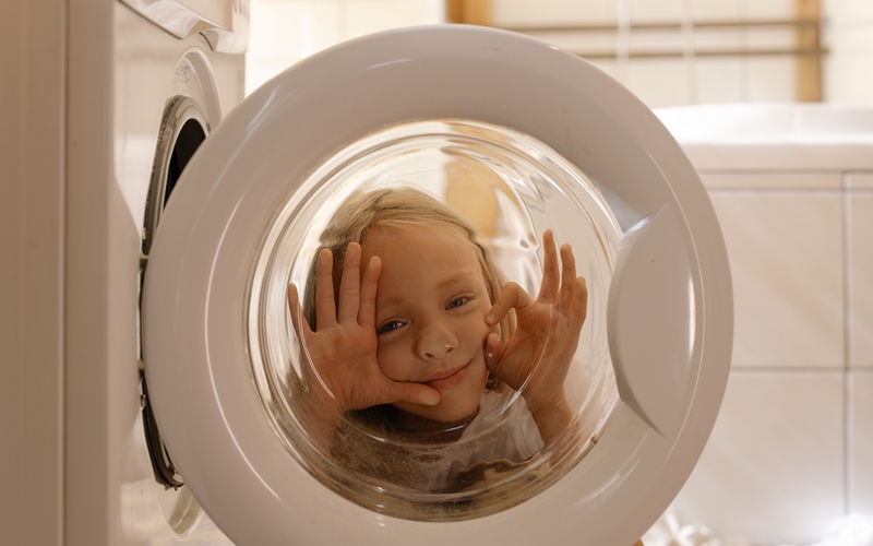 A young girl making a face through the glass of an open washing machine door.