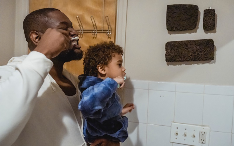 Father and son standing in front of a bathroom mirror brushing their teeth together.