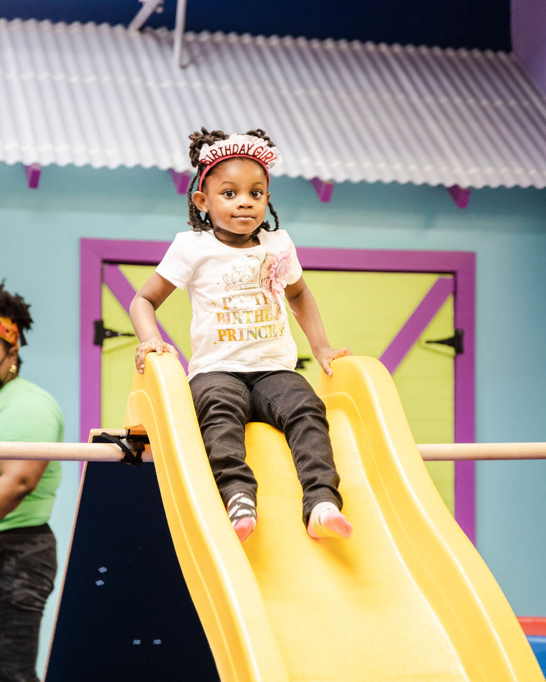 A girl with a birthday girl crown sliding down a yellow slide at Romp n' Roll's kids birthday parties in Midlothian, {fran_state_abbre}.