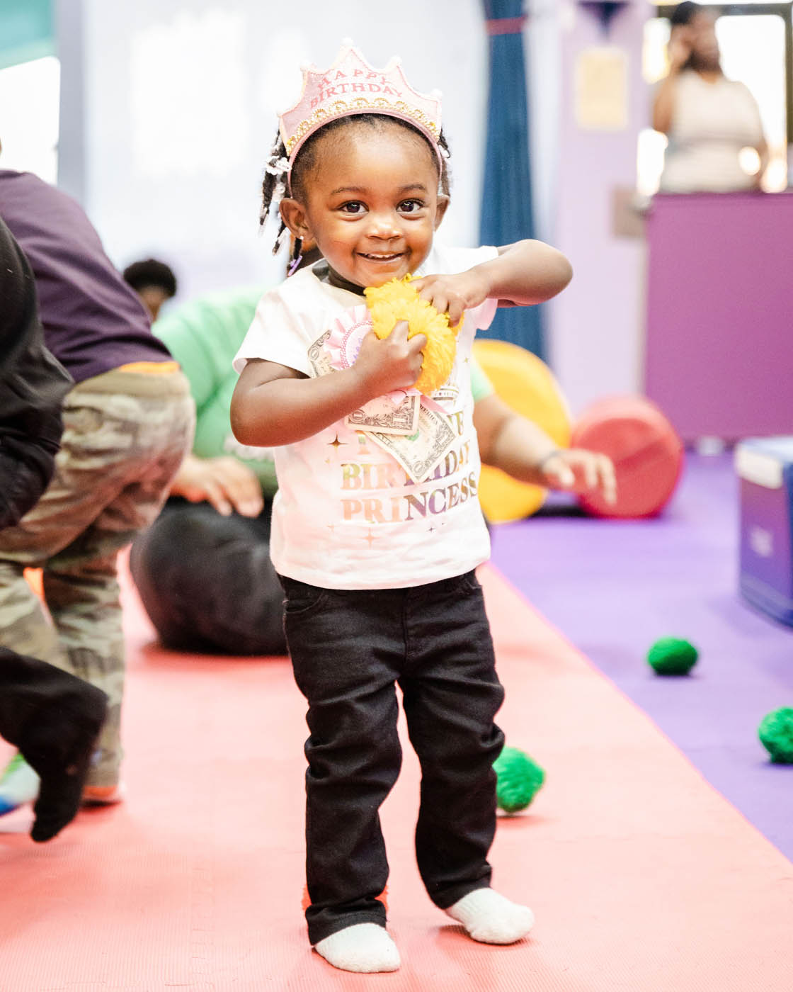 A young girl with a pink princess shirt on holding a yellow fluffy ball - Romp n' Roll children's party places in Glen Allen, VA.