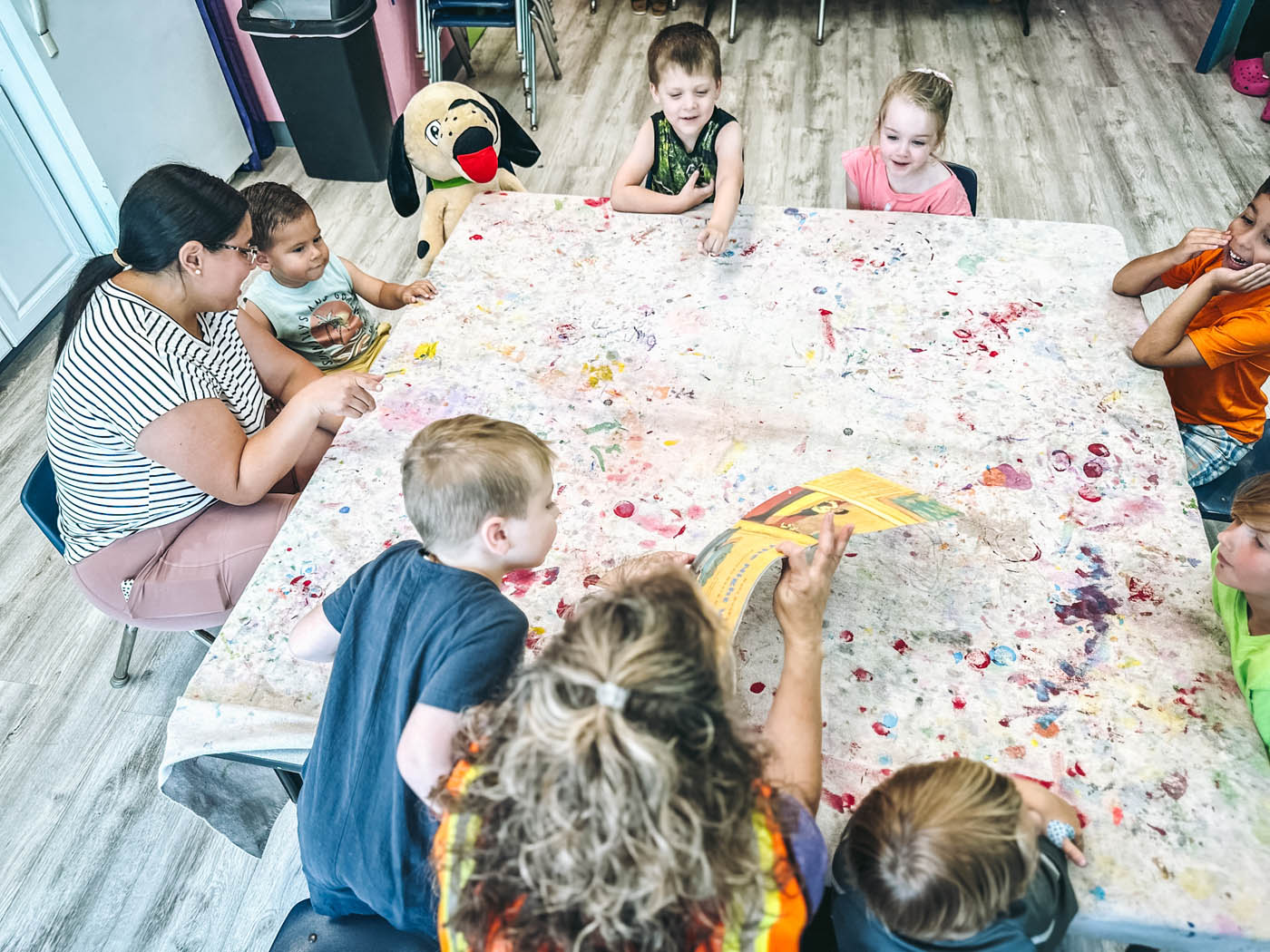 A group of children gathered around a colorful table listening to a Romp n' Roll children's fitness franchise owner read a book out loud.