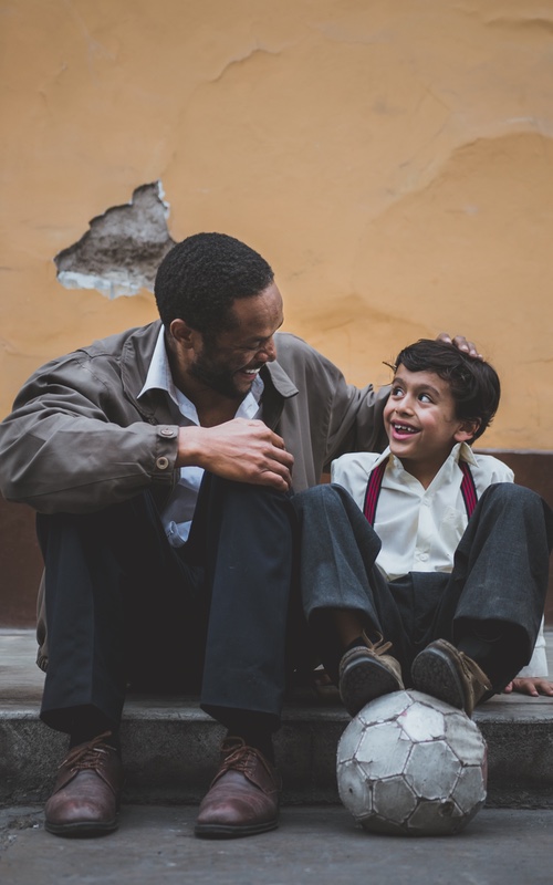 Father and son sitting on a sidewalk curb smiling at each other with a soccer ball.